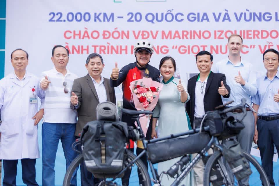 Marino, con un ramo de flores y la bicicleta cargada delante, rodeado de médicos y responsables del Hospital Vinh Duc bajo la pancarta de bienvenida
